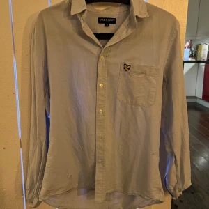 Ljusblå skjorta, Lyle and Scott - Säljer en stilren ljusblå skjorta från Lyle & Scott med klassisk krage och knappstängning. Skjortan har en broderad logotyp på bröstfickan och är långärmad. Perfekt för en avslappnad men ändå uppklädd look på fest eller med familjen. Skjortan har ett litet hål på framsidan. Hör av er om ni har några frågor