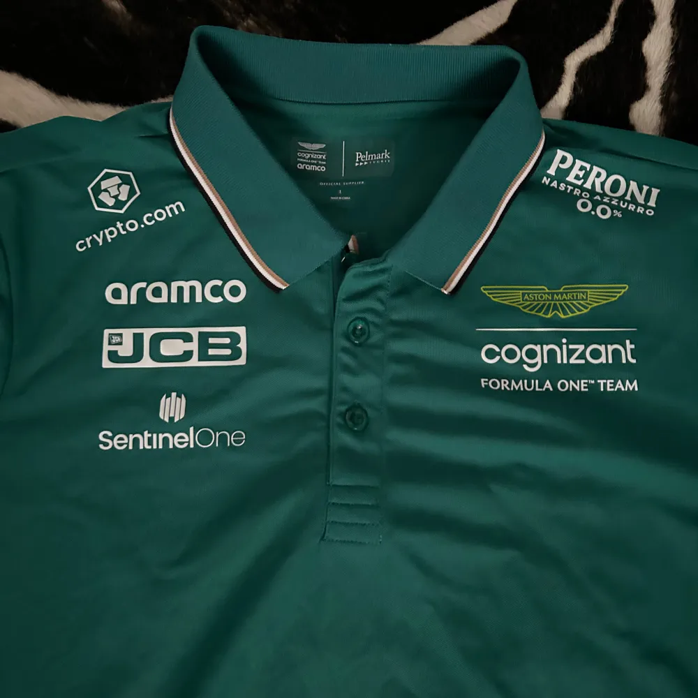 Säljer en snygg grön pikétröja från Aston Martin Cognizant Formula One Team. Tröjan har flera sponsorloggor som JCB, aramco och Peroni. Den har en klassisk krage med knappar och är perfekt för alla F1-fans. Passar både till vardags och för att kolla på F1!. Skjortor.