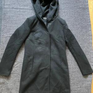 Svart kappa med huva - Säljer en stilren svart kappa från Only. Den har en praktisk huva och stängs med både dragkedja och tryckknappar. Två fickor i sidorna. Perfekt för höst och vinter, med långa ärmar och en skön passform. Aldrig använts. Kappan är i mycket fint skick och redo för nya äventyr! 
