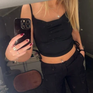 Svart croppad topp med knyt - Snygg svart croppad topp med smala axelband och knyt i sidan. Den är gjord i ett stretchigt silkes-aktigt material. Perfekt för en kväll ute eller en avslappnad dag. Enkel och stilren design som passar till det mesta🖤✨ Nypris 200kr, knappt använd.