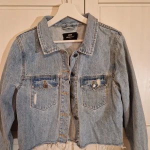 Ljusblå croppad jeansjacka från Dr Denim - Säljer en snygg ljusblå croppad jeansjacka från Dr Denim i strl L. Jackan har en klassisk krage, knappar framtill och två bröstfickor med knappdetaljer.  Jackan är i en något kortare modell, slitenstil och har en rå kant nedtill för en trendig look.