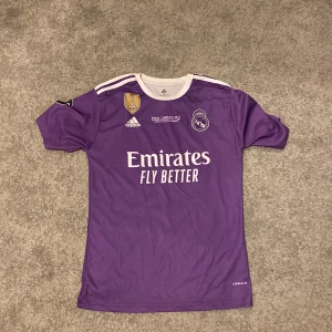 Lila Real Madrid fotbollströja - Säljer en lila Real Madrid fotbollströja från Adidas med 'Emirates Fly Better' tryck på framsidan. Tröjan har korta ärmar och vita detaljer. På baksidan finns 'Ronaldo 7'. Perfekt för fans av laget!