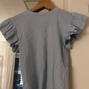Blå  topp med volangärmar - Säljer en söt blå randig topp med volangärmar. Perfekt för en somrig look. Toppen har en normal passform och är tillverkad i ett mjukt material.