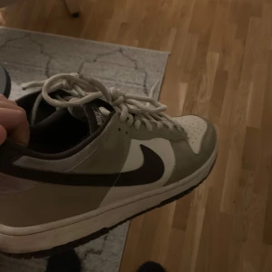 Nike sneakers i beige och svart - Säljer ett par snygga Nike sneakers i beige och svart med klassisk snörning. Perfekta för en stilren look. Skorna har en vit sula och den ikoniska Nike-loggan på sidan.