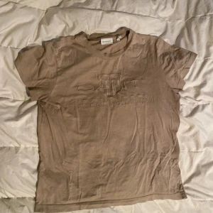 Beige t-shirt från GANT - Säljer en beige t-shirt från GANT med broderad logga på framsidan. T-shirten är kortärmad och tillverkad i bomull, perfekt för en avslappnad stil.