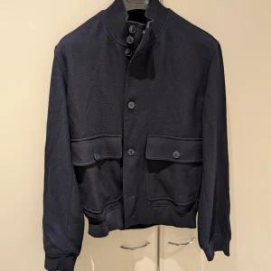 Mörkblå jacka från Massimo Dutti i stl M - Snygg mörkblå jacka från Massimo Dutti med knappar framtill och två stora fickor. Jackan har en ribbad krage och långa ärmar, perfekt för en stilren look.