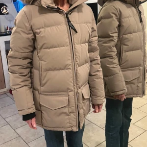 Beige dunjacka från Canada Goose - Säljer en beige dunjacka från Canada Goose med pälskantad huva och dragkedja. Jackan har långa ärmar och praktiska fickor framtill. Perfekt för kalla vinterdagar.