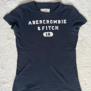 Abercrombie & fitch T-shirt  - Mörkblå, tight T-shirt med korta ärmar från abercrombie & fitch. Trycket sticker ut lite vilket är snyggt! Passar även S