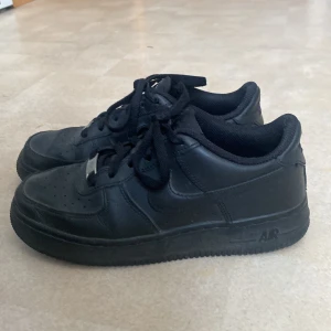Svarta Nike Air Force sneakers - Säljer ett par klassiska svarta Nike Air Force sneakers. Skorna har en stilren design med snörning, använda men i bra skick ändå. Har slitits i hälen på insidan på ena skon, bara tyget som gått sönder men inget som känns. Har crease skydd som jag använt en gång bara som jag kan skicka med om det önskas. 