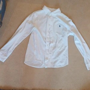 Vit skjorta från Polo Ralph Lauren - Klassisk vit skjorta från Polo Ralph Lauren gjord av bomull med broderad logotyp i blått på bröstet. Skjortan har långa ärmar och knappar framtill. Perfekt för en stilren look. 