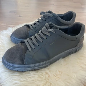 Grå sneakers från Axel Arigato - Snygga grå sneakers från Axel Arigato med stilren design. Skorna har en kombination av skinn och mocka, vilket ger en exklusiv känsla. De har snörning och en robust sula med märkets logotyp präglad på sidan och sulan.