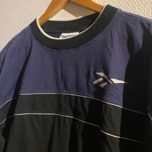 T-Shirt från Reebok - Säljer en cool blå och svart tröja från Reebok med vita detaljer. Tröjan har korta ärmar och Reebok-loggan på bröstet och ärmen. 