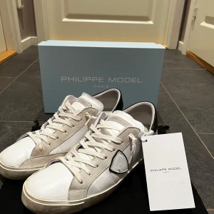 Vita sneakers Philippe Model - Snygga vita sneakers från Philippe Model med svarta detaljer på hälen och en unik logotyp på sidan. Skorna har en klassisk snörning och är tillverkade i skinn och mocka för en stilren look. Skorna är väldigt försiktigt använda och mycket bra skick