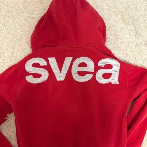 Röd huvtröja från Svea - Snygg röd huvtröja från Svea med dragkedja och stor logga på ryggen. Perfekt för en avslappnad stil med en bekväm passform. Huvan ger extra värme och stil.