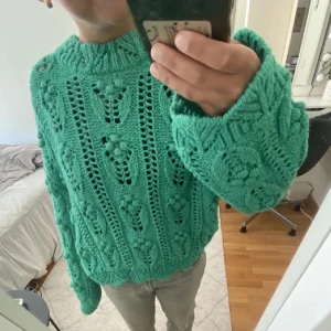 Grön stickad tröja - Säljer en grön stickad tröja med ett unikt hålmönster. Tröjan har långa ärmar och en bekväm passform, perfekt för kyligare dagar. Den är både stilren och mysig, idealisk för att hålla sig varm med stil.