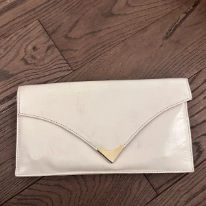 Vit kuvertväska med gulddetalj - Elegant vit kuvertväska med en stilren gulddetalj på framsidan. Väskan har en praktisk magnetknapp och flera fack inuti för enkel organisering. Perfekt för att bära det nödvändigaste med stil.