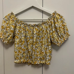 Blommig offshoulder topp från Gina Tricot - Säljer en söt offshoulder topp från Gina Tricot i storlek XS. Toppen har ett blommigt mönster i gult och vitt med små gröna blad. Den har korta ärmar och elastiska kanter för en bekväm passform. Perfekt för sommardagar!