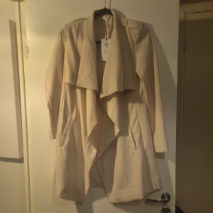 Beige kappa från Object - Snygg beige kappa från Object med en elegant draperad krage och knappdetalj på axeln. Perfekt för att ge en stilren look till din outfit. Passar bra till både vardag och mer uppklädda tillfällen.