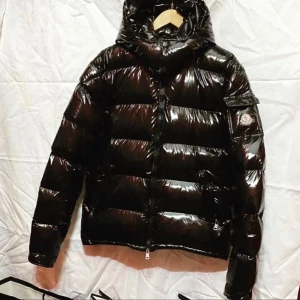 Svart Moncler Maya Jacka Storlek M - Snygg svart pufferjacka från Moncler med glansig finish och huva. Jackan har en dragkedja framtill och långa ärmar. Perfekt för kalla dagar med sin quiltade design som håller värmen.  