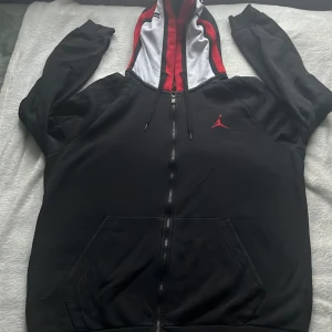 Svart hoodie Jordan - Säljer en svart hoodie från Jordan med en cool röd och vit huva. Tröjan har en dragkedja framtill och en liten röd logga på bröstet. Perfekt för en avslappnad stil.
