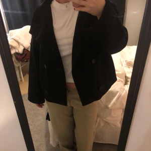 Svart kappa med knappar - Snygg svart kappa med dubbelknäppning och bred krage. Jackan har en lös passform och långa ärmar, perfekt för höst och vinter. En stilren och tidlös design som passar till många tillfällen.