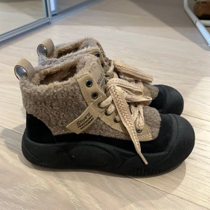 Beige och svarta boots från Male Monkey - Snygga och varma boots från Male Monkey i beige och svart. Skorna har snörning och en mysig pälsdetalj runt skaftet. Perfekta för kyligare dagar med en robust sula för extra grepp.