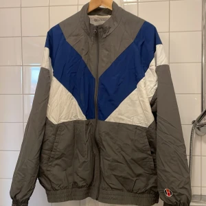 Homebound jacka  - Snygg vindjacka i grått med blå och vita detaljer. Jackan har en dragkedja framtill och elastiska muddar vid ärmslut och nederkant. Perfekt för en sportig look.