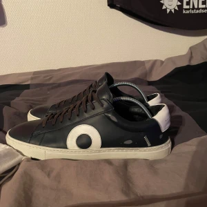 Mörkblå sneakers från Oliver Cabell - Snygga mörkblå sneakers från Oliver Cabell med vita detaljer och snörning. Skorna har en stilren design med en vit sula och en rund vit cirkel på sidan. Perfekta för en avslappnad stil. Kontakta vid frågor. Bara några små skador på ena längst fram men de syns inte när skorna används