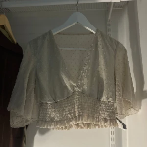Beige blus med volangdetaljer - Säljer en söt beige blus med v-ringning och volangdetaljer. Blusen har korta ärmar och ett smockat midjeparti som ger en fin passform. Perfekt för en somrig look!