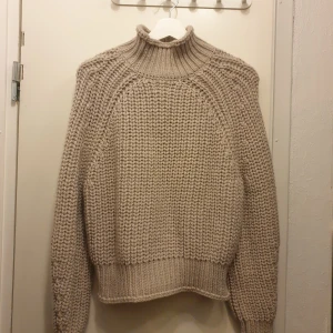 Beige stickad tröja från H&M - Mysig beige stickad tröja från H&M med hög krage och långa ärmar. Perfekt för kyliga dagar med sin tjocka och varma design. Tröjan har en ribbad struktur vid krage, ärmslut och nederkant. Helt ny med lapp kvar. Storlek M