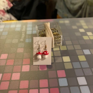Svamphängen örhängen - Charmiga örhängen med svampmotiv i rött och vitt. Perfekta för att ge en lekfull touch till din stil. Hängena är små och lätta, vilket gör dem bekväma att bära hela dagen.