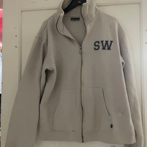 Beige fleecejacka från SOC - Säljer en beige fleecejacka från SOC med dragkedja och broderade bokstäver 'SW' på bröstet. Jackan har långa ärmar och en hög krage för extra värme. Perfekt för kyliga dagar!
