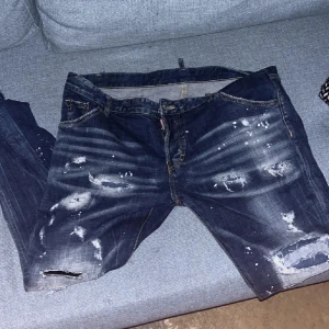 Blå jeans från Dsquared2 - Säljer ett par blå jeans från Dsquared2 med slitna detaljer och hål för en edgy look. Jeansen har en knappgylf och är tillverkade i Italien. Perfekta för dig som vill ha en unik stil. OBS!!! Skinnet på vänster ben är lite löst men går att fixa enkelt med hjälp av textil lim 