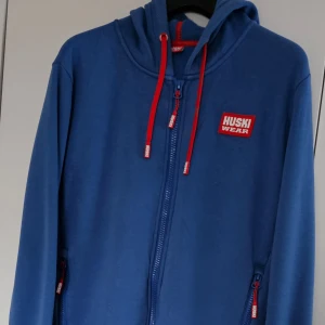 Blå huvtröja från Huski Wear - Snygg blå huvtröja från Huski Wear med röda detaljer. Tröjan har dragkedja framtill och en praktisk huva med röda snören. Perfekt för en avslappnad stil.