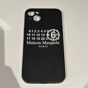  Maison Margiela IPhone 14 Pro Max mobilskal - Snyggt svart mobilskal från Maison Margiela för IPhone 14 Pro Max med deras ikoniska nummerdesign i vitt. Skalet har utskärningar för kamera och knappar, vilket ger enkel åtkomst och skydd. Perfekt för att ge din telefon en stilren look. Har någon liten fett fläck men som bara syns i visst ljus 