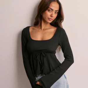 Nelly topp - Trendig svart ribbad topp med lång ärm och knytning i midjan. Perfekt för en stilren look. Helt ny och oanvänd 