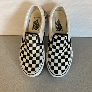 Svartvita rutiga slip-on sneakers från Vans - Säljer ett par klassiska Vans slip-on sneakers i svartvitt rutigt mönster. De är i välanvänt skick men fortfarande supercoola och perfekta för vardagsbruk. Storlek dam 36. Passar perfekt till både jeans och shorts för en avslappnad stil.