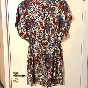 Klänning från KappAhl - Säljer en blommig klänning från KappAhl i storlek M. Den har ett härligt mönster i blått och rött på en vit bakgrund. Klänningen är kort och är justerbar, för att justera midjans bredd. Oanvänd.