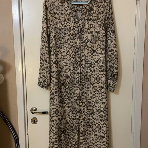 Långklänning från H&M - Säljer en superfin mönstrad långklänning från H&M i storlek M. Klänningen har ett coolt svart och beige mönster och är gjord i ett mjukt material. Den har långa ärmar och ett bälte i midjan. Ganska bra skick