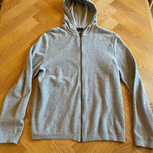 Grå hoodie med dragkedja från Massimo Dutti - Säljer en stilren grå hoodie i mjukt material med dragkedja framtill. Perfekt för både vardag och mysiga stunder. Den har en klassisk passform och långa ärmar. Huvan ger extra värme och stil. Passar till alla tillfällen! 