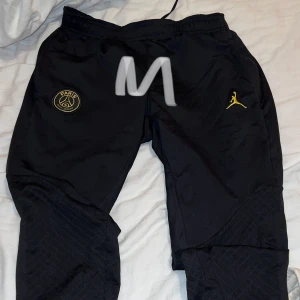 Svarta träningsbyxor från Jordan x PSG - Säljer ett par svarta träningsbyxor från Jordan x PSG. Byxorna har elastisk midja och är dekorerade med PSG-loggan och den ikoniska Jumpman i gult. Perfekta för fotbollsträning eller chill. De är i bra skick och redo för nya äventyr!