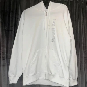 Vit hoodie med dragkedja - Säljer en stilren vit hoodie med dragkedja framtill. Den har en bekväm passform och är perfekt för vardagsbruk. Det finns en snygg detalj med glittriga stenar längs dragkedjan. Hoodien har långa ärmar och en klassisk känguruficka. Perfekt för både höst och vår!