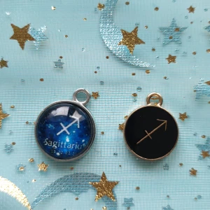 Sagittarius berlocker i silver och guld - Två snygga berlocker med stjärntecknet Skytten. Den ena har en blå bakgrund med stjärnor och texten 'Sagittarius' i silver, medan den andra har en svart bakgrund med en gyllene pil. Perfekta för att lägga till en personlig touch till ditt smycke!
