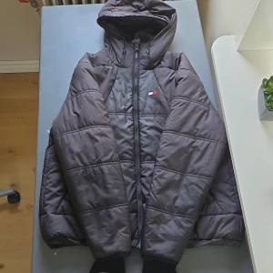 Svart pufferjacka från Tommy Hilfiger - Säljer en svart pufferjacka från Tommy Hilfiger. Den har en sportig look med huva och dragkedja framtill. Jackan är perfekt för kyligare dagar och har en quiltad design som ger extra värme. Storlek M och är en sample-modell, vilket gör den unik