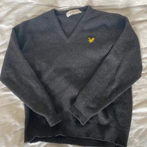 Säljer en snygg och klassisk mörkgrå tröja från Lyle & Scott. Den är gjord av 100% ren lammull vilket gör den supermjuk och varm. Tröjan har en v-ringad hals och långa ärmar, perfekt för kyliga dagar. Den har också en liten gul logga på bröstet som ger en stilren touch.