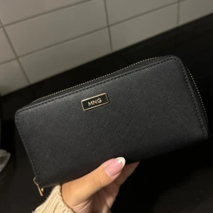 Svart plånbok från MNG - Säljer en stilren svart plånbok från Mango. Den har en praktisk dragkedja och flera fack inuti för kort och kontanter. Perfekt för att hålla ordning på dina saker!