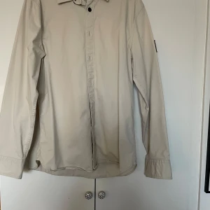 Beige skjorta från Calvin Klein Jeans - Säljer en stilren beige skjorta från Calvin Klein Jeans i storlek L. Skjortan har långa ärmar och en klassisk krage. Den är i regular fit och har en diskret logga på ärmen. Perfekt för både vardag och fest! Nypris 999kr