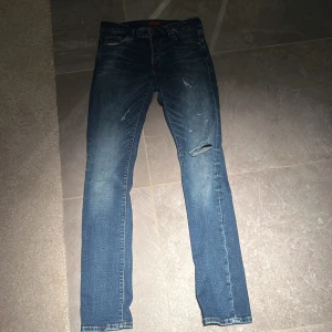 Jack & Jones jeans - Säljer ett par snygga blå jeans med coola slitningar på framsidan. De har en klassisk femficksdesign och en bekväm passform. Perfekta för en avslappnad stil!