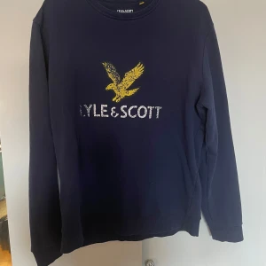 Mörkblå tröja från Lyle & Scott - Säljer en snygg mörkblå tröja från Lyle & Scott med deras ikoniska gula örnlogga på bröstet. Tröjan är i storlek L och har långa ärmar. Perfekt för en casual look eller när du vill ha något bekvämt men stilrent. Passar bra till jeans eller chinos!