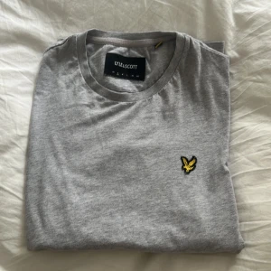 Grå t-shirt från Lyle & Scott - Säljer en stilren grå t-shirt från Lyle & Scott. Den är kortärmad och har en liten broderad logga på bröstet i gult och svart. Perfekt för en avslappnad stil!